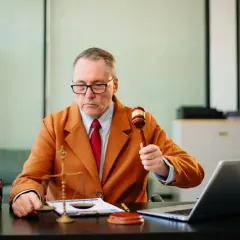 Sędzia w pomarańczowej marynarce trzyma młotek, gotowy do ogłoszenia wyroku. Na biurku waga sprawiedliwości i laptop.