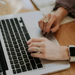 Kobieta pisze na laptopie, analizując odsetki ustawowe. Na palcach ma obrączkę i pierścionek, a na nadgarstku smartwatch.