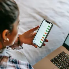 Kobieta sprawdza na telefonie wykresy giełdowe, zastanawiając się, ile można zarobić na giełdzie. Obok leży laptop z mapą inwestycji.