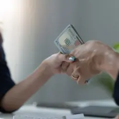 Dwie dłonie przekazują sobie plik banknotów. Jedna z nich ma złoty pierścionek. To symboliczne przedstawienie zawarcia umowy pożyczki.