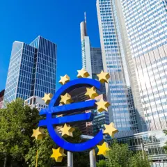 Symbol euro przed nowoczesnymi wieżowcami, siedzibą europejskiego banku centralnego.