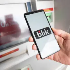 Dłoń trzyma smartfon z logo Blik przed bankomatem. Sprawdź, ile można wypłacić z bankomatu Peako.