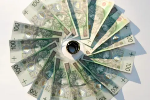 Złote banknoty o nominale 100 zł ułożone w okrąg, z pierścieniem pośrodku. Oficjalny sposób na oszczędzanie.