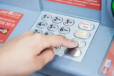 Dłoń wpisuje PIN na bankomacie, obok karta płatnicza. Może to oznaczać próbę sprawdzenia salda lub wypłaty, która doprowadzi do debetu na koncie.