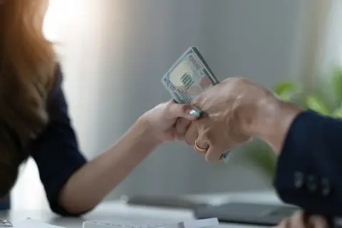 Dwie dłonie przekazują sobie plik banknotów. Jedna z nich ma złoty pierścionek. To symboliczne przedstawienie zawarcia umowy pożyczki.