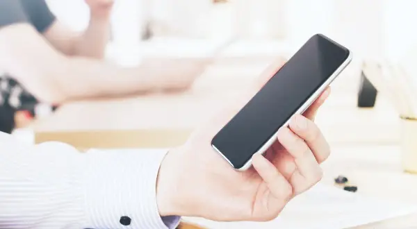 Dłoń trzyma smartfon, być może przeglądając ofertę kredyt orange. W tle widać biurko i inne osoby.