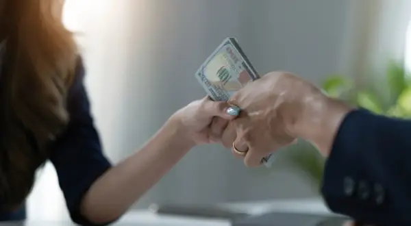 Dwie dłonie przekazują sobie plik banknotów. Jedna z nich ma złoty pierścionek. To symboliczne przedstawienie zawarcia umowy pożyczki.