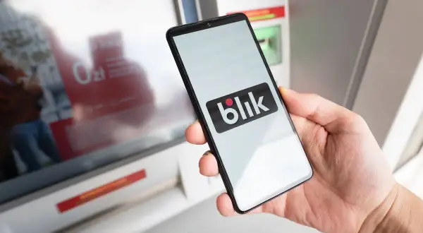 Dłoń trzyma smartfon z logo Blik przed bankomatem. Sprawdź, ile można wypłacić z bankomatu Peako.
