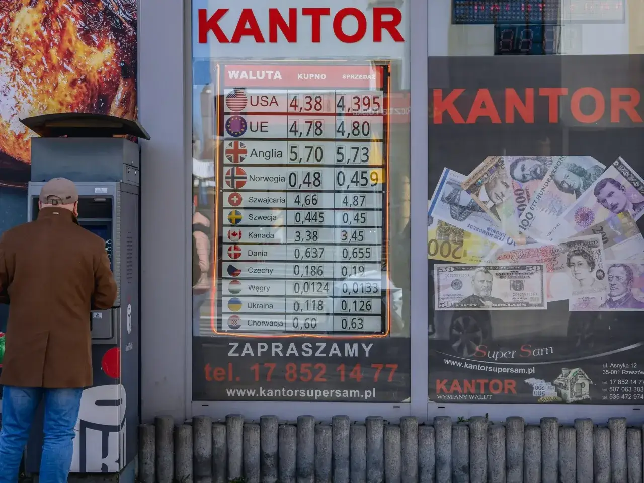 Kantor wymiany walut z cennikiem i banknotami. Mężczyzna korzysta z bankomatu obok.