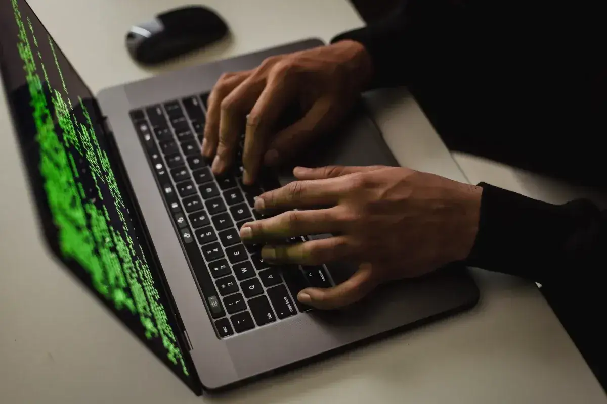 Dłonie piszą kod na laptopie, ekran wyświetla zielony tekst. To obraz cyberataku, gdzie haker pracuje w ukryciu.