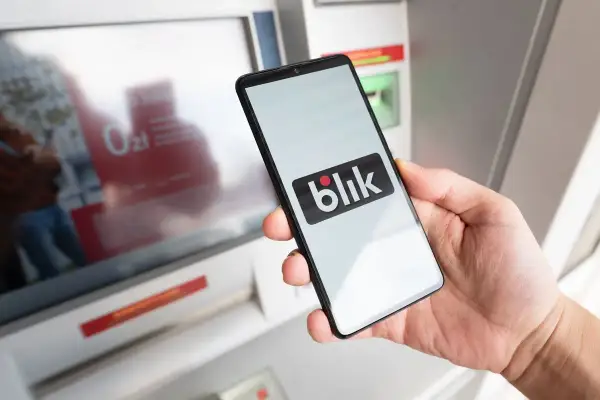 Dłoń trzyma smartfon z logo Blik przed bankomatem. Sprawdź, ile można wypłacić z bankomatu Peako.
